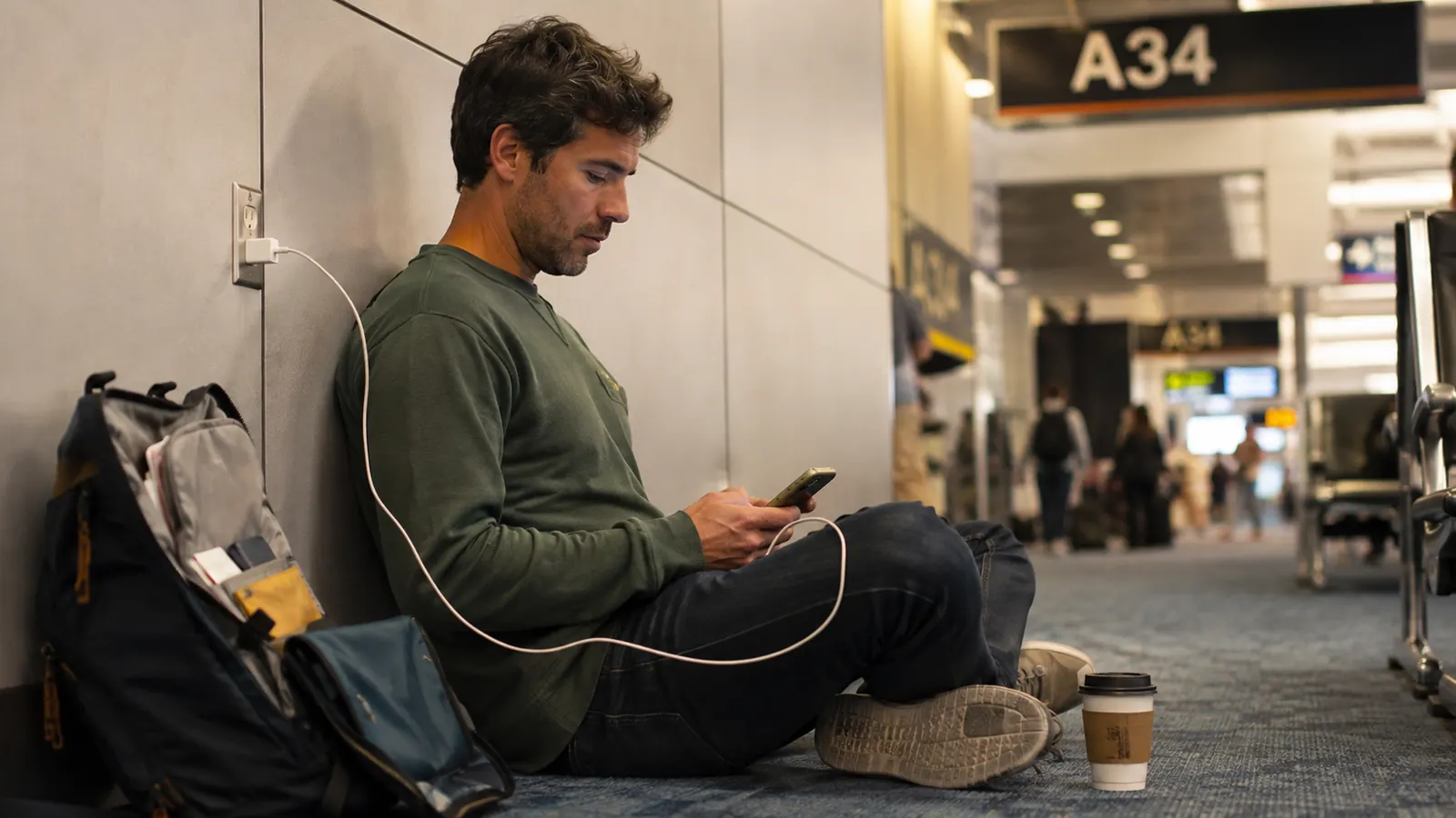 Traveler sitting on the floor of a crowded airport terminal near a boarding gate, phone plugged into a small power bank while checking a boarding pass