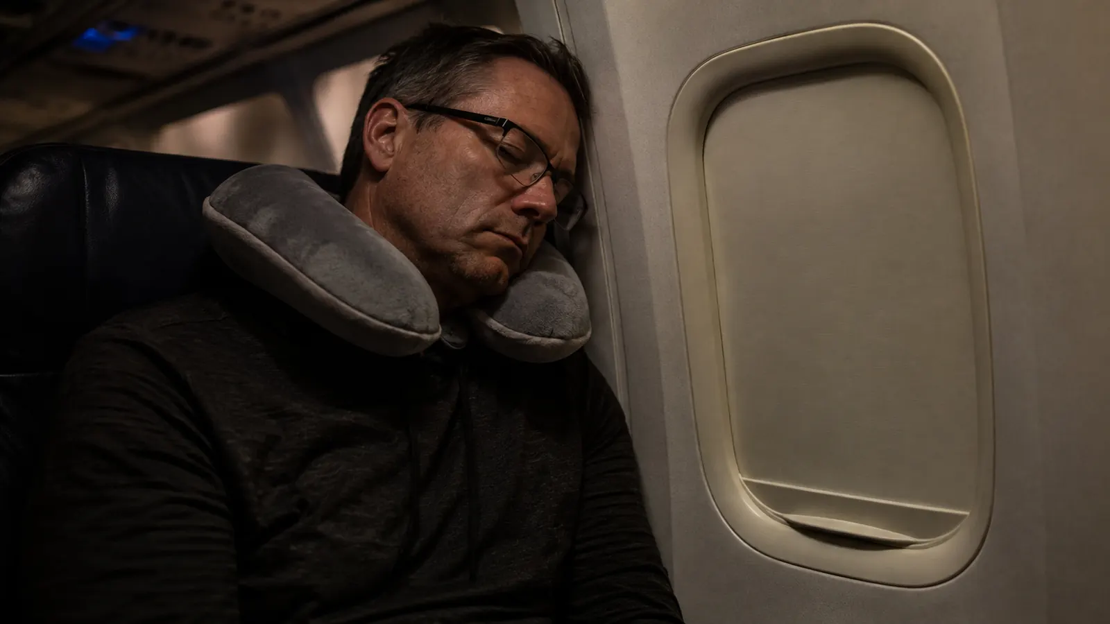 Traveler asleep in an airplane window seat at night wearing a curved neck pillow, cabin lights dimmed