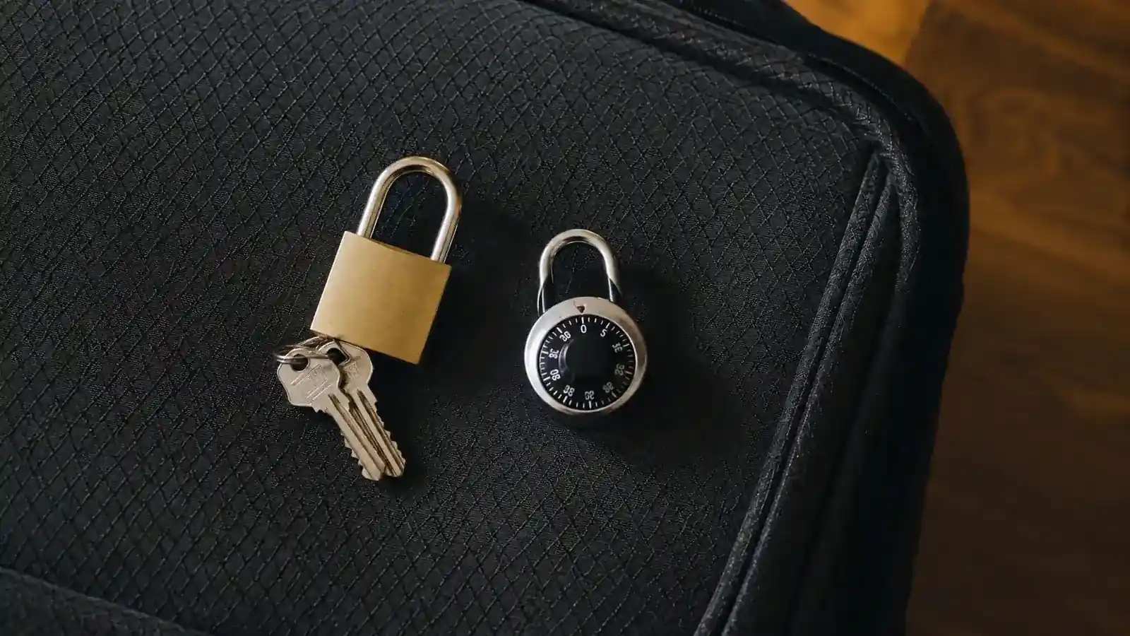 A keyed brass-colored TSA luggage lock and a small combination dial lock resting side by side on a suitcase next to a set of keys