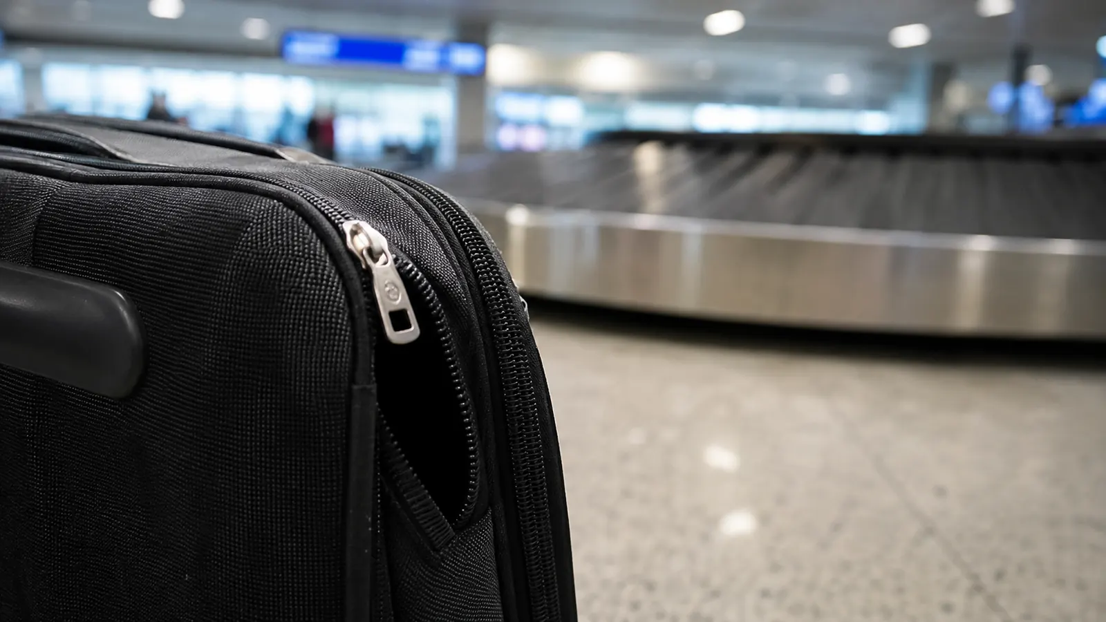 A worn zipper pull on a suitcase hanging slightly open at an airport carousel